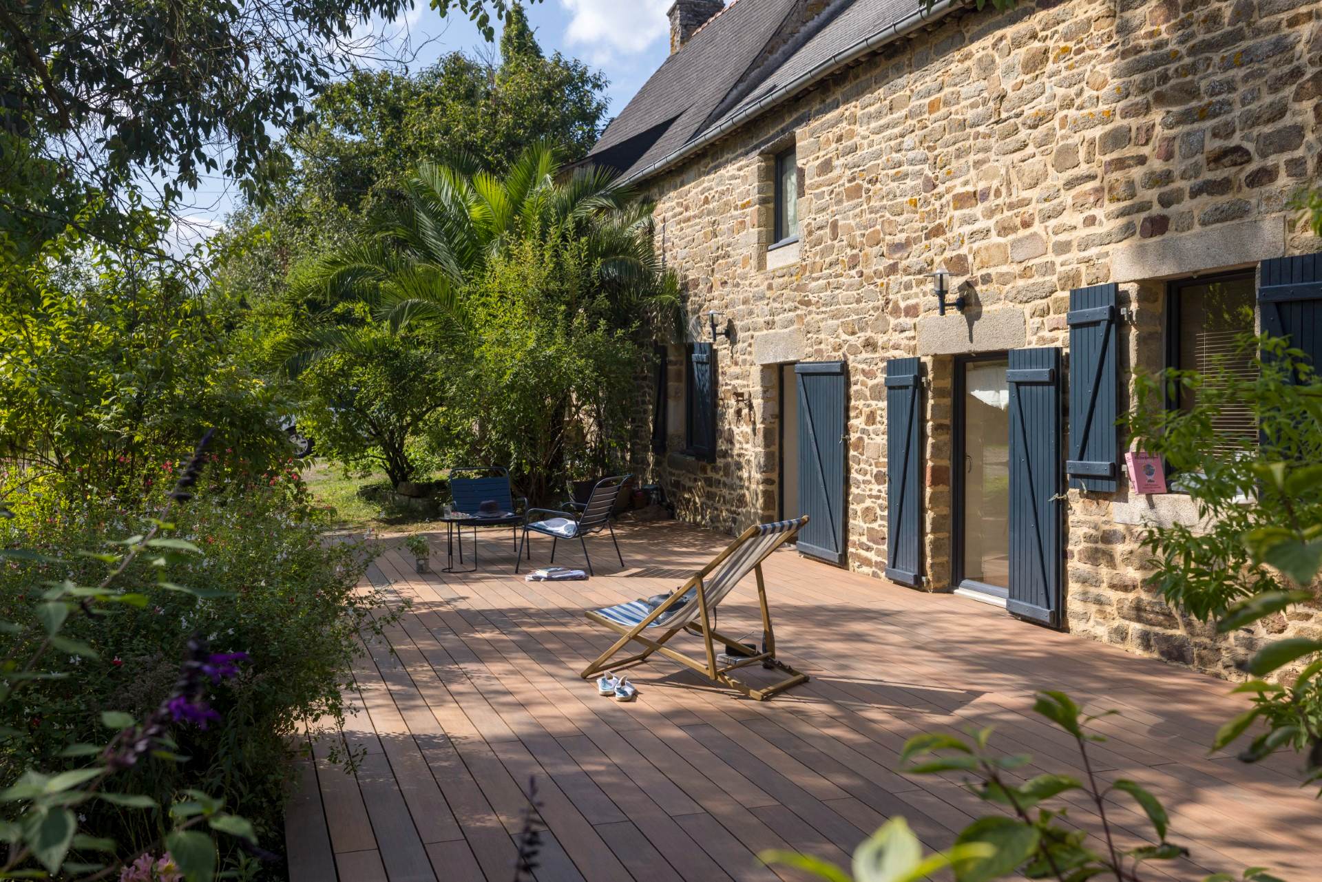 Grande terrasse en lames de bois composite teinte ipé entourant une maison bretonne en pierre avec volets bleus, transat et salon de jardin, végétation luxuriante, parfait exemple d'aménagement extérieur avec bois composite pour terrasse, alternative au bardage traditionnel