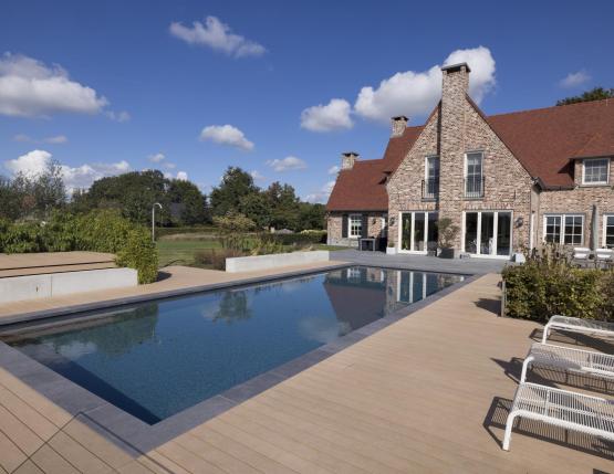 Élégante maison en pierre avec toit rouge, terrasse en bois composite entourant une piscine rectangulaire, mobilier extérieur moderne et jardin paysager en arrière-plan