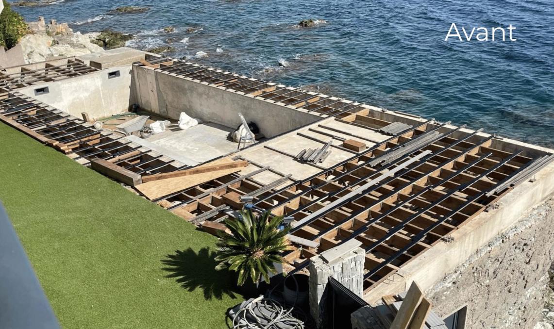 Structure en cours de construction 'Avant' l'installation, montrant l'ossature en bois d'une terrasse en bord de mer avec vue panoramique sur l'eau. La préparation du support pour recevoir les lambourdes en aluminium réversible Réversil de Silvadec. Un palmier est visible au premier plan près de la fondation en béton.
